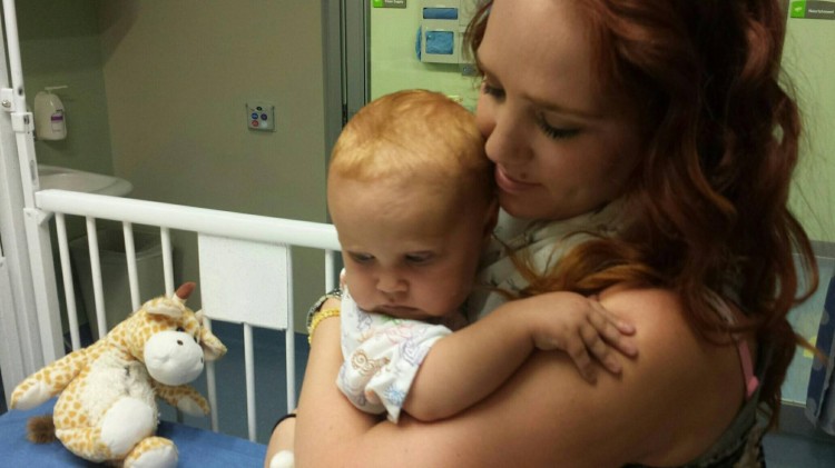 A mom holds a baby next to a crib in a hospital. 