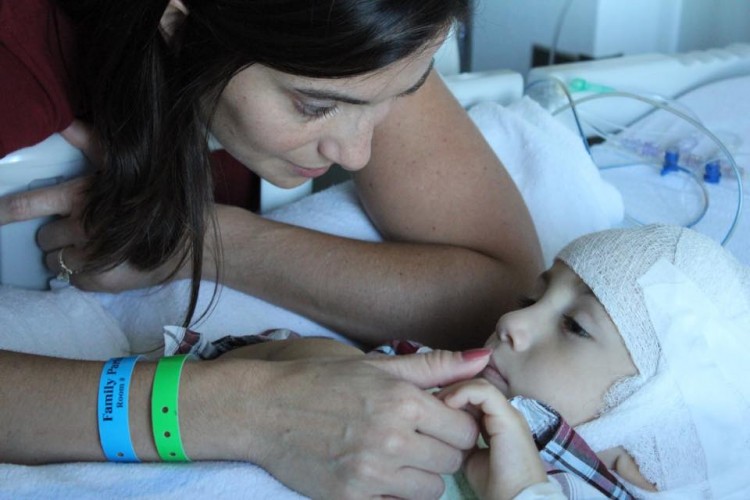 A mom gently puts her thumb on the mouth of a little boy, who's laying in a hospital bed. 