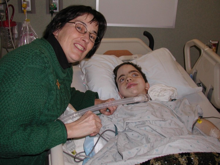 A woman smiles next to her son, who lays in a hospital bed. 