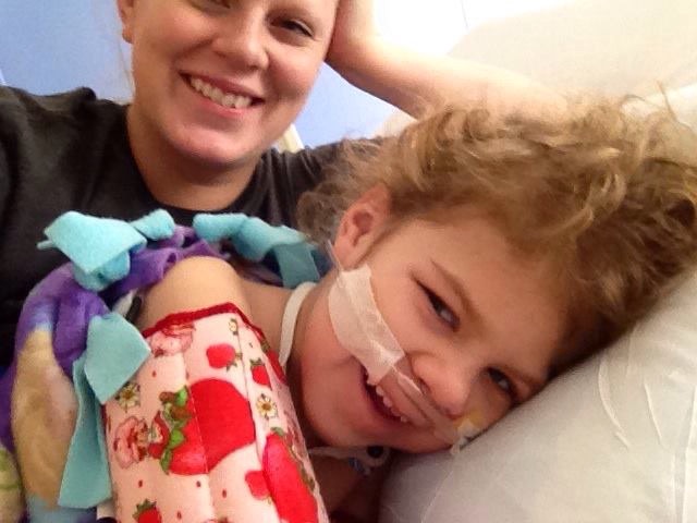 A mom smiles with her daughter, who's laying in bed with a breathing tube. 