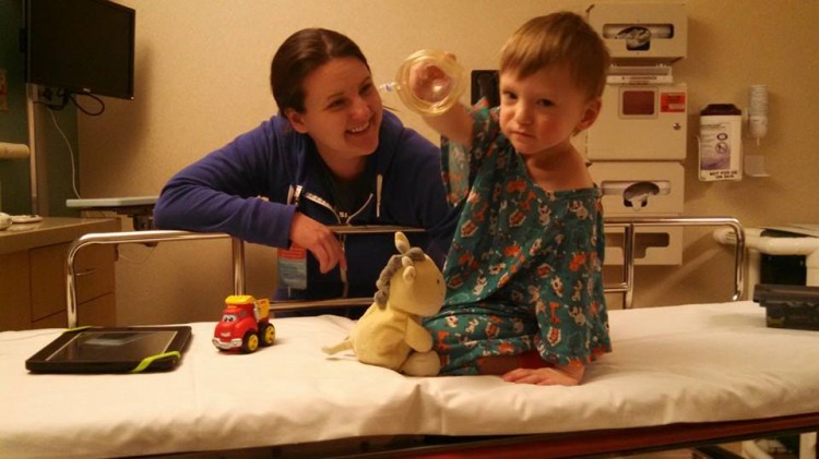 A mom smiles at her son, who sits in a hospital bed with toys. 