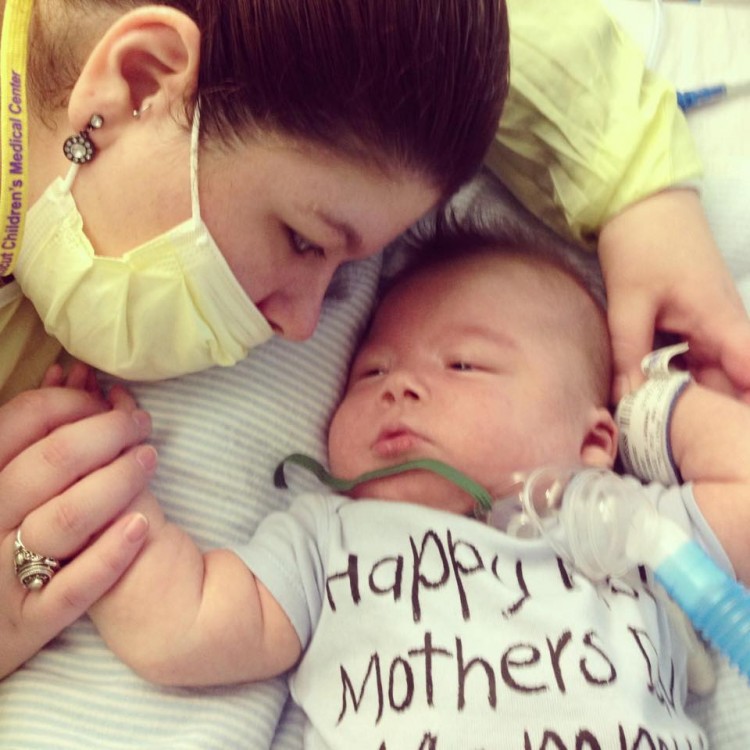 A mom with a face mask leans over her baby. The baby's shirt says Happy Mother's Day.