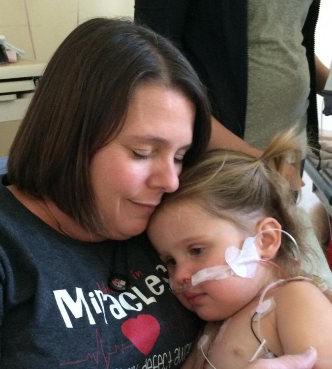 A little girl with a breathing tube leans her head on her mother's shoulder.