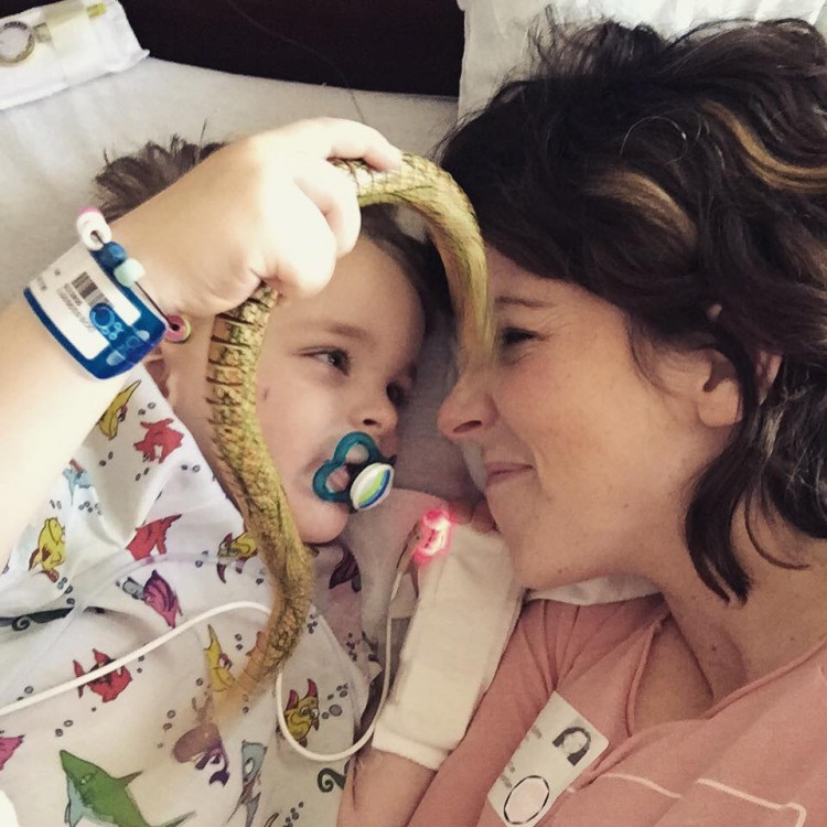 A mom and her son look at each other while a laying in a hospital bed. The boy holds a toy snake. 
