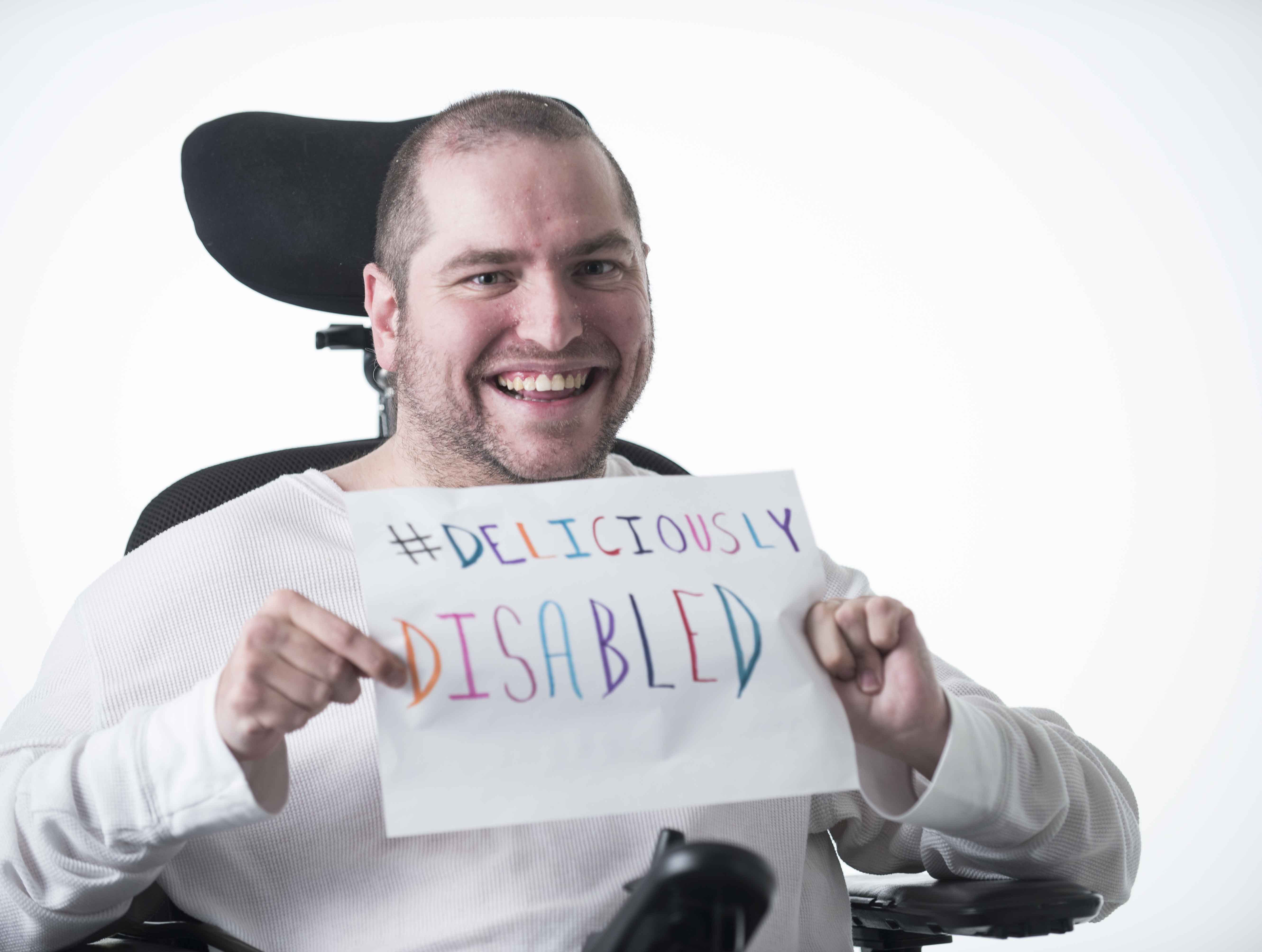 Andrew sits in a wheelchair holding up a sign that says #DeliciouslyDisabled