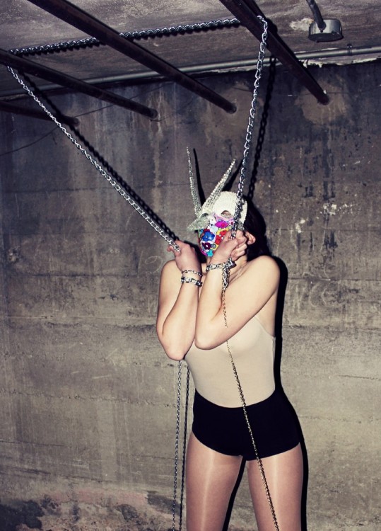 Stones hands are chained to the ceiling. She wears a colorful mask, a beige shirt and black shorts. 