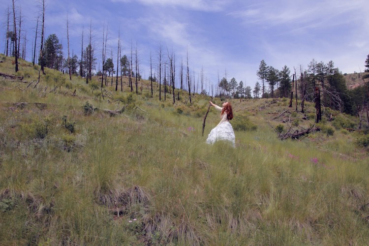 A woman with red hair stands in a bare field. She holds a walking stick and looks upwards.
