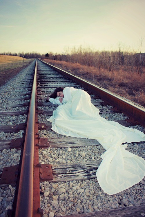 Stone lays on train tracks in a white gown. 