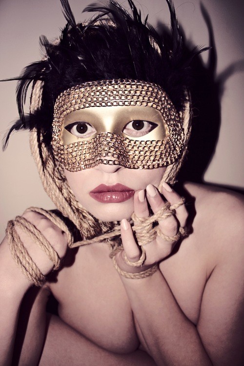 Stone stares into the camera, wearing a golden mask. Her hands are tied together with rope. 