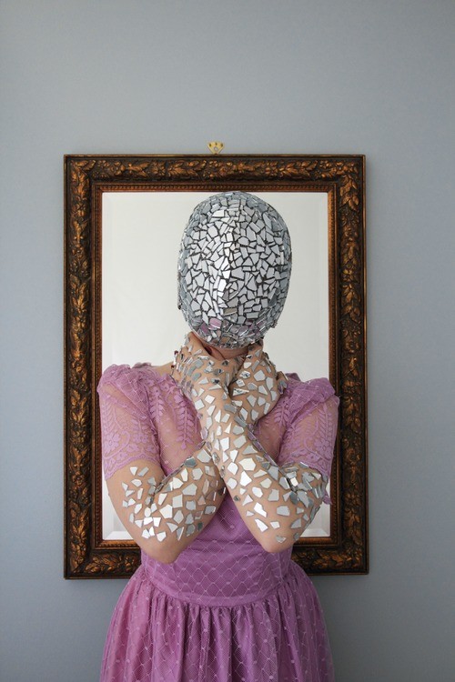 Stone wears a pink dress and a mask made of sharps of glass. She also has glass adorning her arms. She stands in front of an empty picture frame. 