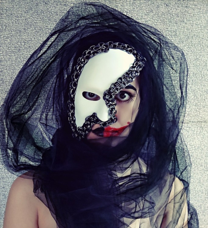 Stone stares into the camera wearing a white mask covering half her fact. The side of her face without a mask shows thickly painted red lipstick. 