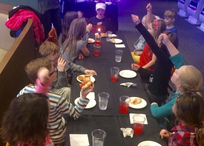 9-year-old boy having a birthday party -- he and his friends are sitting at a table, eating pizza and cheering as he starts to blow out the candle on his cupcake