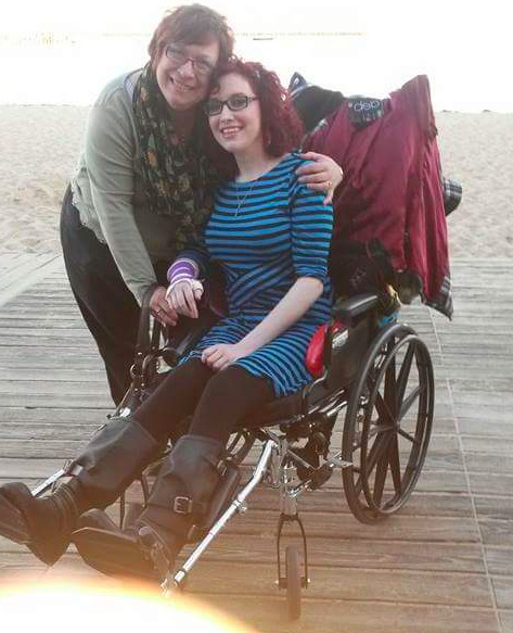 Two women on the beach. One woman is in a wheelchair, and the other woman is standing next to her and has her arm wrapped around her. Their heads are touching and they are smiling.