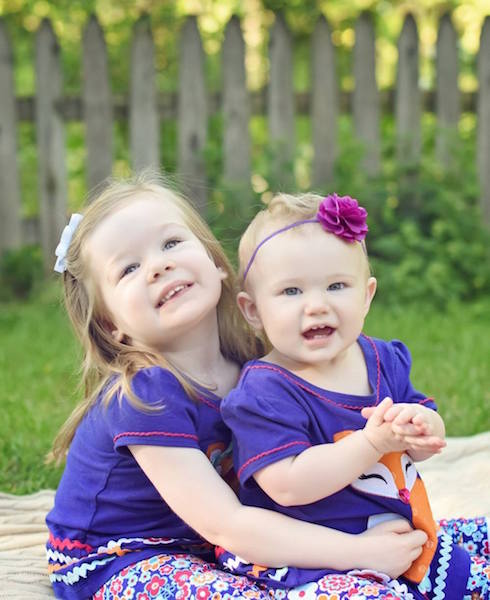 two young sisters wearing matching purple shirts and flower headbands posing together in their yard