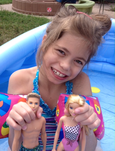 young girl holding up two barbie dolls as she sits in inflatable pool