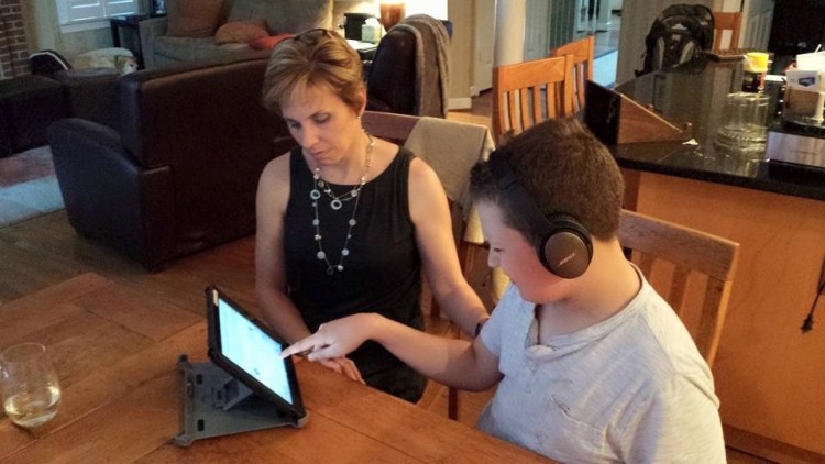 mother sits with her nonverbal son as he writes with the help of a tablet