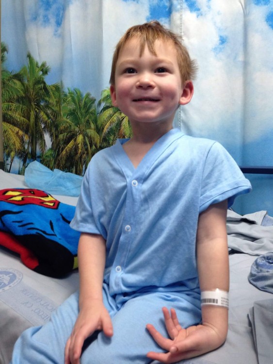 Jack sitting on a hospital bed with a beach-themed curtain behind him and a superman towel.