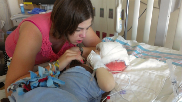 Kathy looking at her son, who lays in a hospital bed. His head is covered with gauze.
