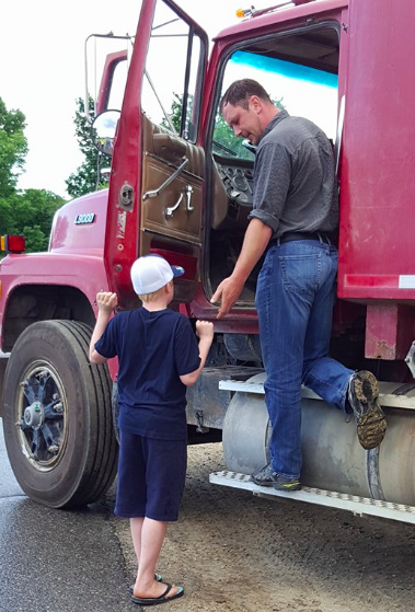 A man standing near the door of a red construction truck, holding his hand out to the author's son