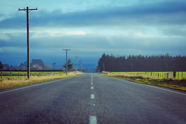 a road with a cloudy sky in the distance, stretching through fields