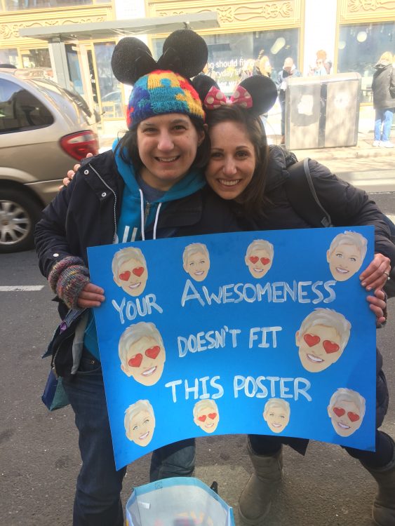 Two women dressed as Mickey and Minnie Mouse holding a sign for Ellen Degeneres Show that says [your awesomeness doesn't fit this poster] with photos of Ellen on it