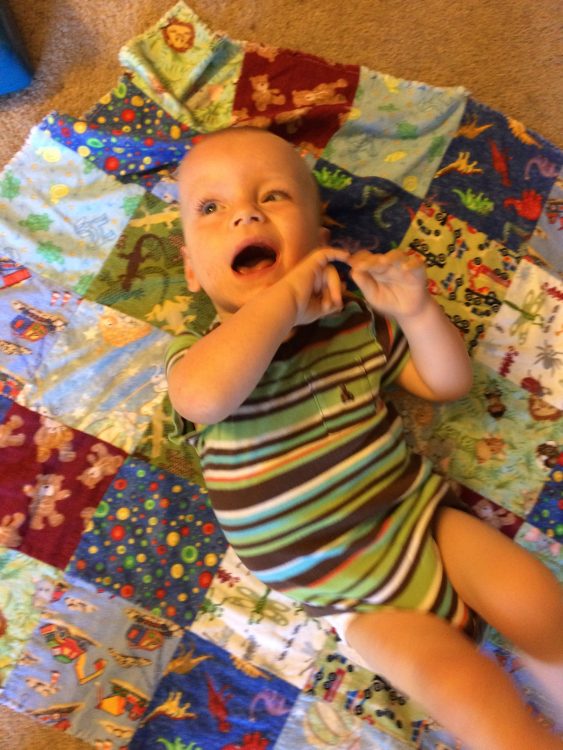 Baby laying on a quilt, smiling, and wearing a blue-green-brown stripped onsie