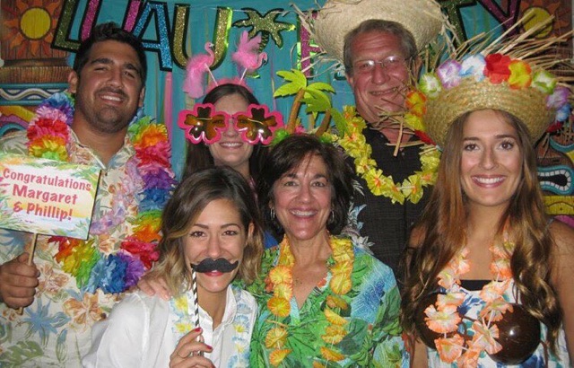 A group of 5 people in a photo booth wearing silly hats, over-sized glasses, in a Hawaiian themed background