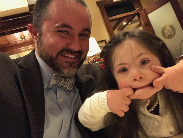 Dad posing with daughter with Down syndrome while she makes a silly face at the camera
