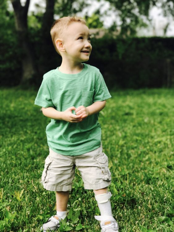 Little boy standing on his backyard, wearing a green shirt, tan shorts, white socks, white leg braces, and white shoes. he is looking to the side, smiling.