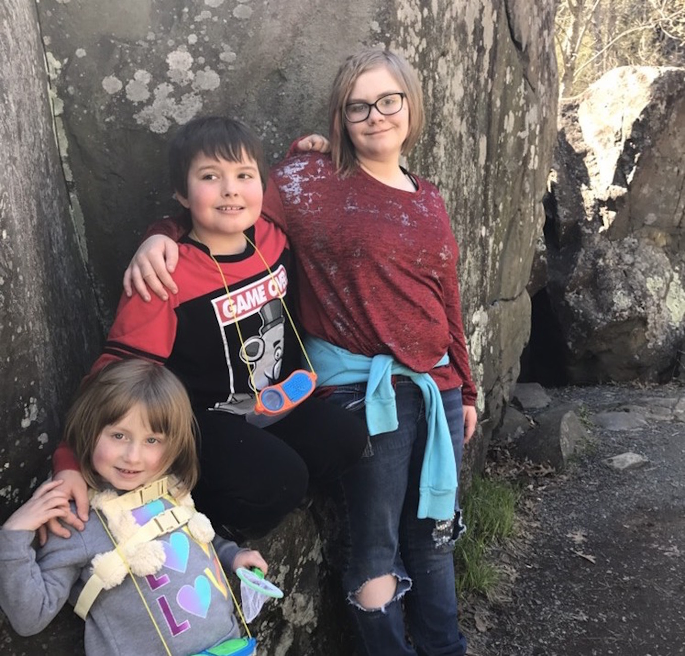 three children with arms around each other, standing in front of boulder outdoors
