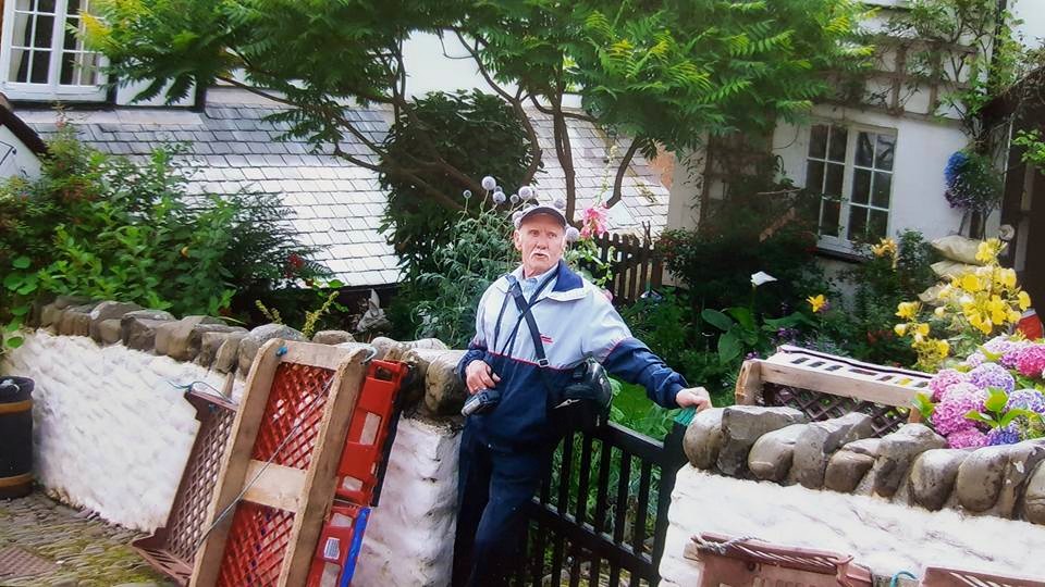 image of elderly man in front of old cottage holding camcorder, pulling a silly face