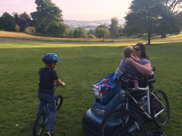 woman in power wheelchair outside with boy on bike and holding young child