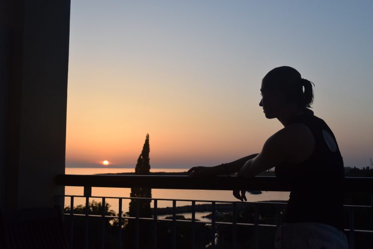 silhouette of a woman standing on a balcony overlooking a lake at sunset