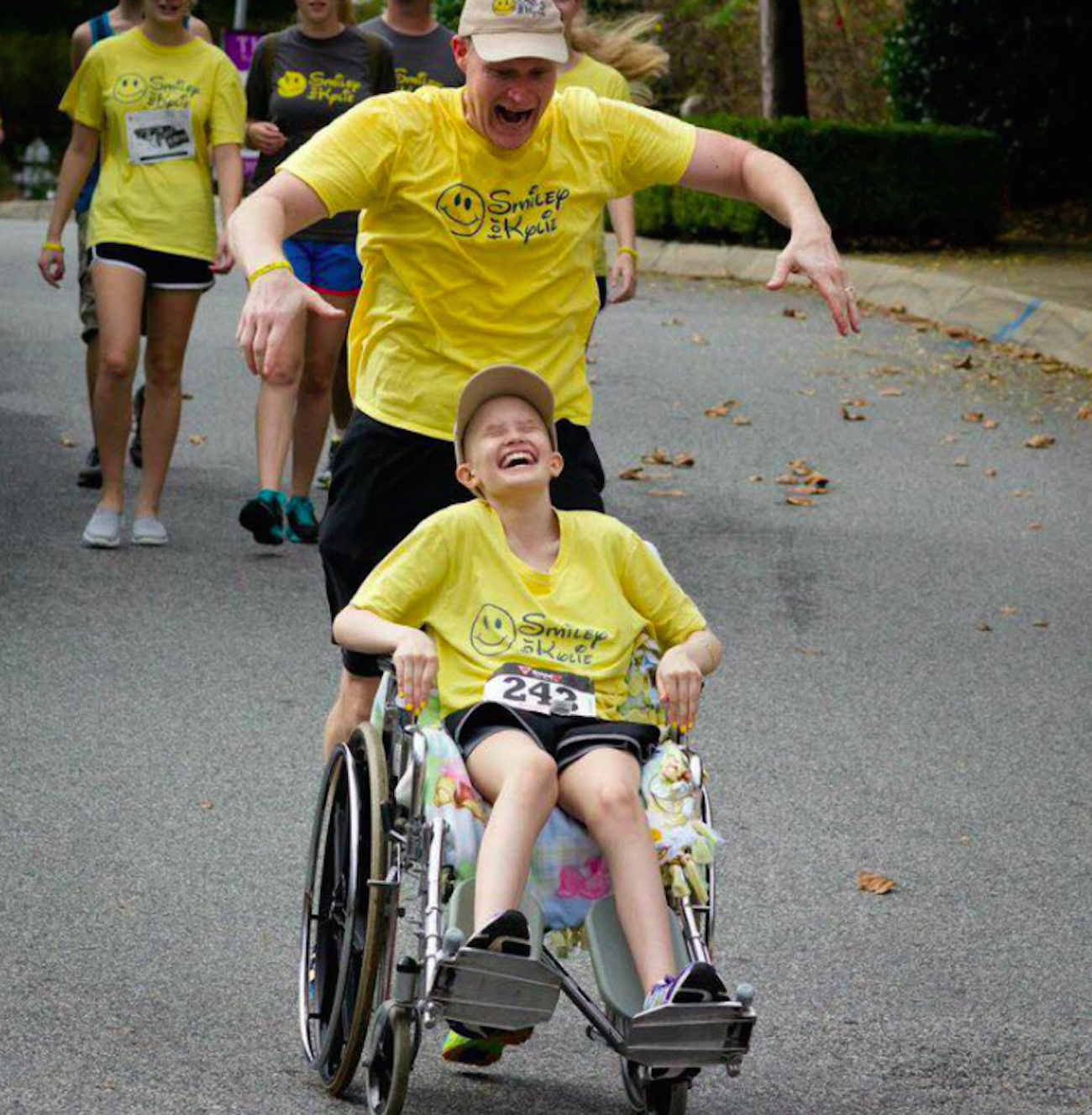 the author pushing his daughter in a wheelchair, both of them laughing together and wearing Smiley for Kylie t-shirts