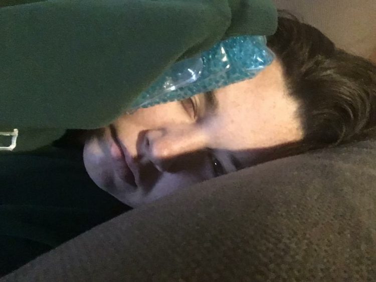 woman lying in bed under a blanket with an ice pack on her head due to a migraine