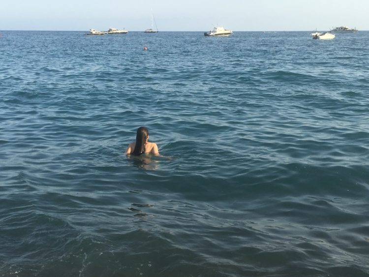 A woman swimming in the ocean, looking at some ships in the distance.