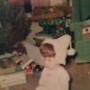 A little boy in all white and a white winter hat sitting by presents under the tree