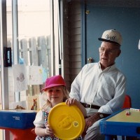 A small girl plays with toys with her grandfather behind her.