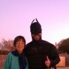 An older, petite woman with glasses, short brown hair and a scarf stands next to her adult son dressed in a batman costume
