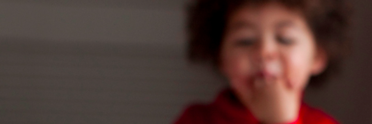 Little boy with hand in his mouth out of focus in the background with chocolate cake in front