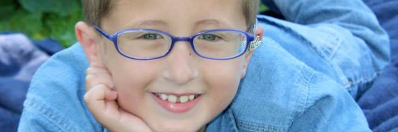 A boy with glasses with one hand on his cheek looking up and smiling into camera