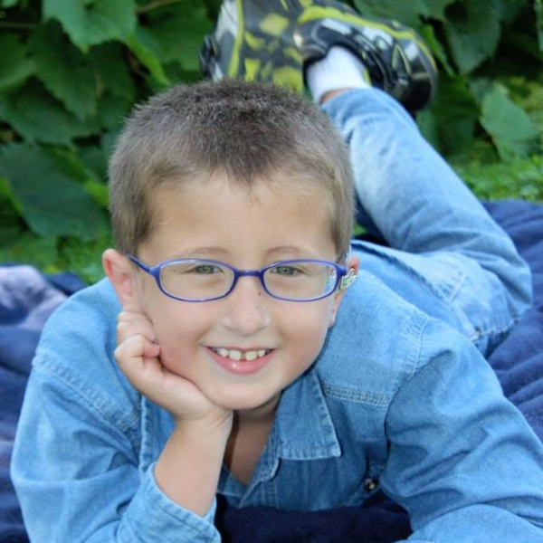 A boy with glasses with one hand on his cheek looking up and smiling into camera