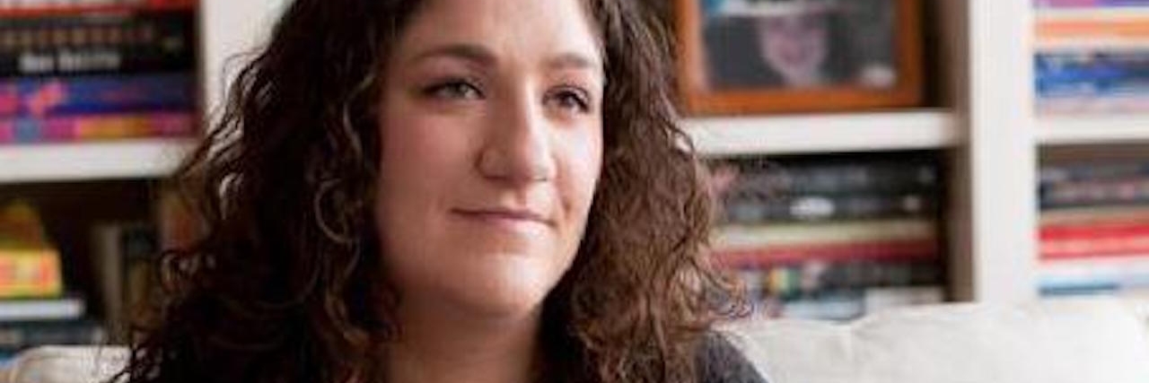 Hyper IGE Syndrome: Why I No Longer Keep My Disease a Secret A young woman with long, dark, curly hair in all black sits on a couch and looks off into the distance