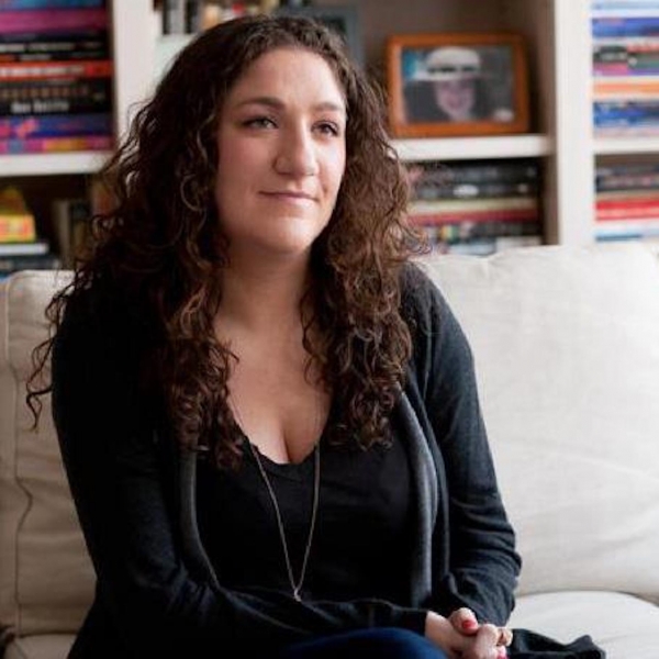 A young woman with long, dark, curly hair in all black sits on a couch and looks off into the distance
