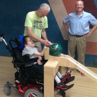 A man stands next to a young man in a wheelchair as he helps adjust a bowling ball on a ramp in front of him