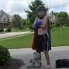 A shirtless boy with a cap stands outside on a sidewalk with a teddy bear against his foot