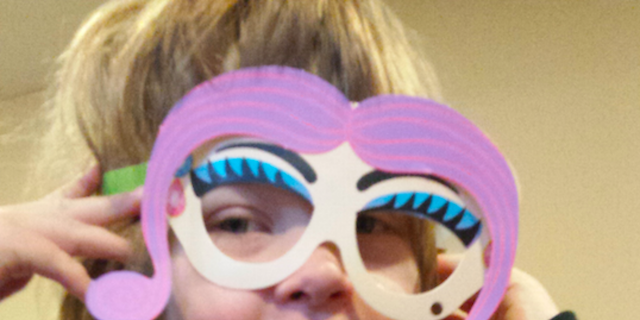 A blond, little girl holding a cutout of fake eyelashes and eyelids up to her face