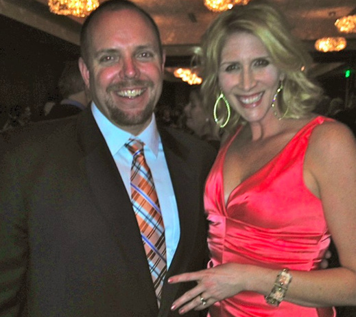 The author and her husband with their arms around each other, smiling for the camera. He's wearing a suit and tie, and she is wearing a red dress and gold earrings.