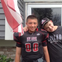 two boys, one in a football jersey, smiling and standing outside their house