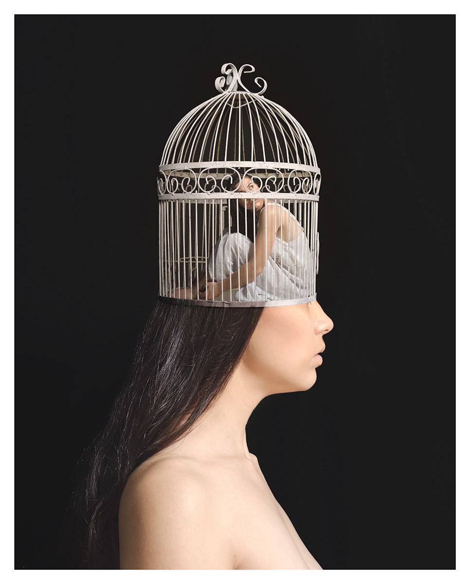 A woman has a bird cage on her head, where another image of the same woman sits, trapped.